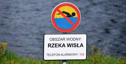 Znak zakazujący kąpieli z tabliczką informacyjną, na której jest tekst: obszar wodny – rzeka Wisła, telefon alarmowy 112. W tle nieostre fale płynącej rzeki.  