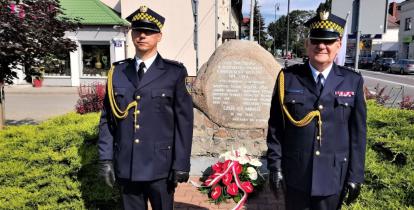 Dwóch strażników miejskich, ubranych w stroje galowe, stoi na baczność obok pomnika upamiętniającego bohaterów Bitwy Warszawskiej z 1920 roku. Pod pomnikiem zostały złożone biało czerwone kwiaty.