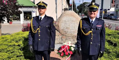 Dwóch strażników miejskich, ubranych w stroje galowe, stoi na baczność obok pomnika upamiętniającego bohaterów Bitwy Warszawskiej z 1920 roku. Pod pomnikiem zostały złożone biało czerwone kwiaty.