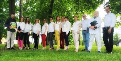 Grupa trzynastu osób. Dziewięć kobiet i czterech mężczyzn. Stoją oni obok siebie. W tle widać drzewa.