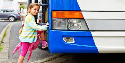 Dziewczynka ubrana w różową sukienkę i zielony sweter wsiada do biało-niebieskiego autobusu.
