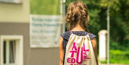 Kilkuletnia dziewczynka idzie po trawie. Na plecach ma jasny worek z napisem kultura, edukacja i zabawa. 