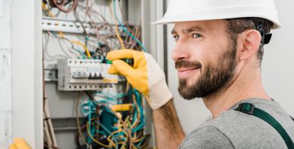 Pomieszczenie gospodarcze, na ścianie zamontowana jest skrzynka elektryczna. Po prawej stronie stoi mężczyzna - elektryk, który sprawdza bezpieczniki. 