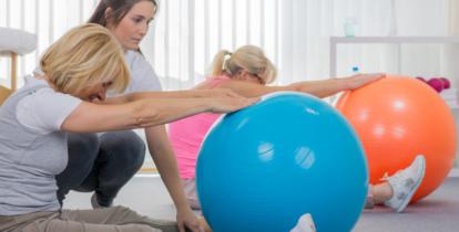 Sala fitness. Dwie kobiety siedzą na podłodze, przed sobą mają duże kolorowe piłki. Wykonują ćwiczenia. Między nimi stoi instruktorka. 