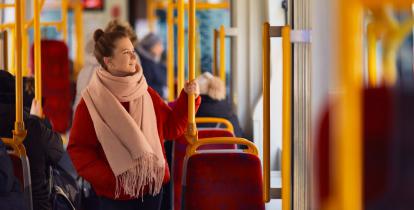 Dorosła kobieta ubrana w jasną kurtkę i szalik na szyi stoi w autobusie. Trzyma się żółtej barierki. 