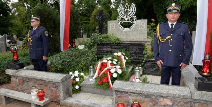 Cmentarz. Przy pomniku ofiar drugiej wojny światowej stoi dwóch mężczyzn w średnim wieku. Ubrani są w mundury Straży Miejskiej. Na pomniku leżą cztery wieńce z biało-czerwonymi kwiatami. Przed pomnikiem stoją cztery znicze, trzy czerwone i jeden biały. Po obu stronach pomnika na sztandarze wiszą biało-czerwone flagi. 