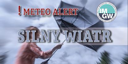 Na górze czerwony napis meteo alert poprzedzony wykrzyknikiem. Obok niego logo Instytutu Meteorologii i Gospodarki Wodnej - niebieski okrąg z białymi literami IMGW, poniżej szary napis silny wiatr. W tle postać z rozwianymi włosami próbująca utrzymać wygięty w górę parasol.  