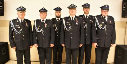 6 mężczyzn ubranych w galowe mundury strażackie stoi obok siebie w rzędzie. W tle widać kremową ścianę budynku. 
