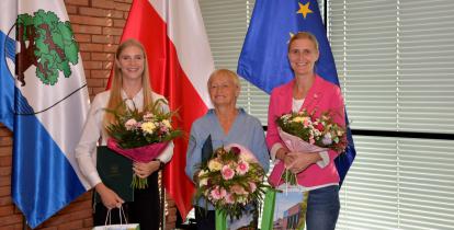 Trzy osoby stoją obok siebie w rzędzie – młoda dziewczyna z długimi blond włosami, kobieta w średnim wieku oraz młoda kobieta. Każda w rękach trzyma bukiet kolorowych kwiatów i torbę prezentową ze zdjęciem Urzędu Miasta i napisem Konstancin-Jeziorna.