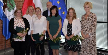  Sala konferencyjna. Siedem osób stoi obok siebie w rzędzie – sześć kobiet i jeden mężczyzna. Cztery kobiety w rękach trzymają dyplomy i białe róże. W tle trzy flagi: Konstancina-Jeziorny, Polski i Unii Europejskiej.