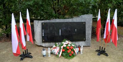 Park. Pomnik ofiar katyńskich. Przy nim stoją znicze oraz leżą: biało-czerwone kwiaty i wiązanka z białymi kwiatami. Po obu stronach pomnika stoją na stojaku trzy biało-czerwone flagi. W tle zielone drzewa.