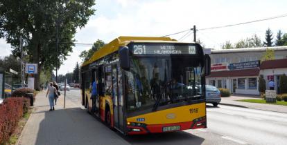 Ulica. Przy przystanku obok chodnika stoi żółto-czerwony autobus komunikacji miejskiej. Z przodu na górze jest napis 251 Metro Wilanowska. Do autobusu wsiada mężczyzna. Chodnikiem idą dwie kobiety.