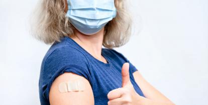 Młoda kobieta stoi na białym tle. Na twarzy ma maseczkę ochronną. Na jej ramieniu przyklejony jest plaster, drugą ręką pokazuje podniesionego kciuka. Widoczna jest od pasa w górę.