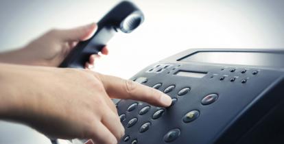 Stacjonarny aparata telefoniczny. Widoczne są dłonie dorosłej osoby, która prawą ręką naciska przyciski z cyframi, a w lewej trzyma słuchawkę. 