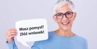 Na zdjęciu uśmiechnięta kobieta – seniorka (krótkie siwe włosy, czarne okulary, ubrana jest w niebieską bluzkę), kobieta trzyma tabliczkę, z napisem: w pierwszym wierszu: Masz pomysł? w drugim wierszu: Złóż wniosek!