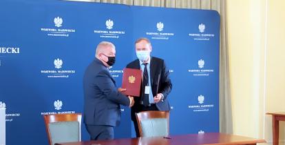 Dwóch mężczyzn stoi naprzeciwko siebie. Ściskaj sobie dłonie. Jeden z nich trzyma w ręku bordową teczkę. Przed nimi stoi duży brązowy stół. 