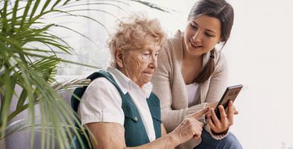 Pokój. Na fotelu siedzi seniorka, obok siedzi młoda dziewczyna. Dziewczyna w ręku trzyma telefon. Seniorka pokazuje palcem coś w telefonie.