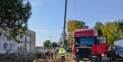 Plac budowy. Po lewej stronie jest wykopany dół, pod budowę fundamentów. Obok niego stoi prostokątny budynek. Po prawej stronie stoi czerwony tir. Za nim widać ramię żurawia budowlanego. 