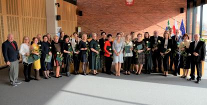 Sala konferencyjna. Dwadzieścia sześć osób stoi obok siebie w rzędzie – sześciu mężczyzn i dwadzieścia kobiet. Większość w rękach trzyma dyplom w zielonej teczce i białą różę.