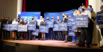 Na scenie stoi grupa osób (kobiety i mężczyźni) w maseczkach trzymających w rękach symboliczne czeki, na których jest tekst: Cyfrowa Gmina, nazwa gminy i 100 tys. zł.