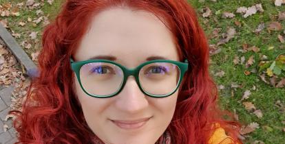 Portret dorosłej kobiety. Ma długie rude włosy, niebieskie okulary. Ubrana jest w żółtą bluzkę. 