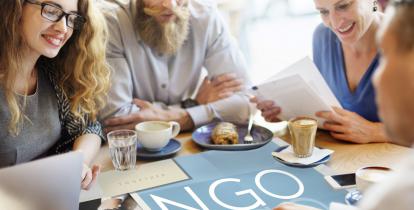 Dwóch mężczyzn i dwie kobiety siedzą przy stoliku, na którym stoją filiżanki z kawę i talerz z ciastkiem. Na środku blatu znajduje się duża niebieska plansza z napisem NGO. 