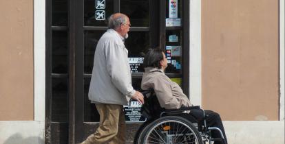 Miasto. Po chodniku idzie mężczyzna, który pcha wózek inwalidzki. Na wózku siedzi kobieta. W tle drzwi budynku. 