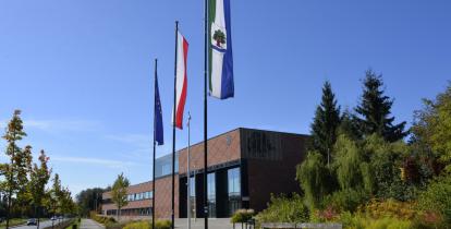 Budynek Urzędu Miasta i Gminy Konstancin-Jeziorna wykonany z czerwonej cegły, przed budynkiem stoją trzy maszty z flagami: Polski, Unii Europejskiej i Konstancina-Jeziorny. Po prawej stronie drzewa i krzewy, po lewej – droga z samochodami. 