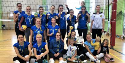 15 kobiet, siatkarek stoi w trzech rzędach na sali gimnastycznej. Ubrane są w niebieskie koszulki. Każda ma złoty medal na szyi. Jedna trzyma w ręku duży puchar. 