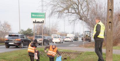 Dwie osoby w kamizelkach odblaskowych z workami zbiera odpady leżące na poboczu drogi. Trzecia osoba, również w kamizelce odblaskowej, nadzoruje ich pracę.