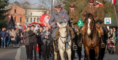 Przemarsz ulicą. Na pierwszym planie trzech mężczyzn w mundurach jadą na koniach, za nimi maszeruje orkiestra dęta oraz ludzie. 