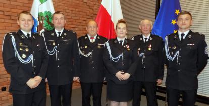 Sześć osób (pięciu mężczyzn i kobieta) w mundurach strażackich stoją w rzędzie na tle flag Polski, Konstancina-Jeziorny i Unii Europejskiej.