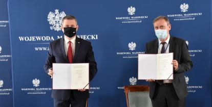 Dwóch mężczyzn stoi obok siebie. W rękach trzymają otwarte teczki. Przed nimi stoi duży brązowy stół.