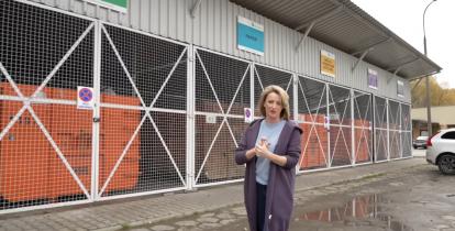 Kobieta stoi na chodniku. W tle jest zadaszony budynek. W środku budynku stoją duże pomarańczowe śmietniki. Na górze do ściany przyczepione są tabliczki z napisami: szkło, papier, metale i tworzywa sztuczne.