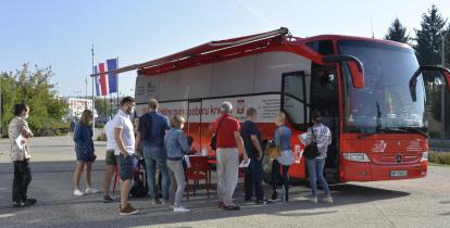 Miasto. Na chodniku stoi czerwony autokar, z boku jest duży napis Mobilny punkt poboru krwi, obok jest godło Polski i napis Minister Zdrowia. Przed autokarem w kolejce stoi dziesięć osób.