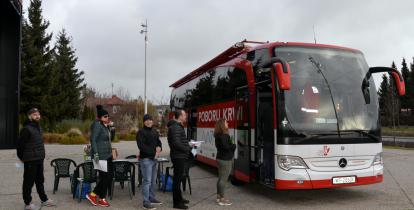 Na chodniku stoi czerwony autokar, z boku jest duży napis Mobilny punkt poboru krwi, obok jest godło Polski i napis Minister Zdrowia. Przed autokarem w kolejce stoi pięć osób.