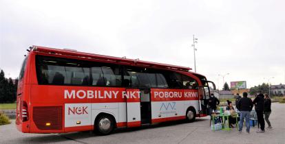 Na chodniku stoi duży czerwony autobus z białym napisem z boku Mobilny Punkt Poboru Krwi. Przed autobusem stoi sześć osób. 