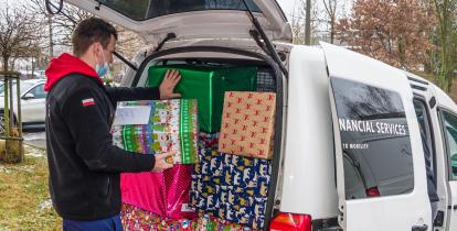 Samochód dostawczy, który ma otwarty bagażnik. Znajdują się w nim duże pudła – prezenty świąteczne. Obok samochodu stoi mężczyzn, który pakuje do niego kolejną paczkę.