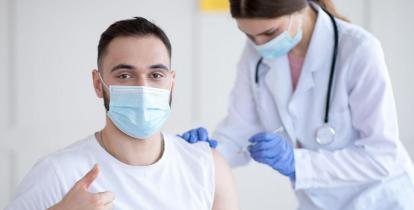 W gabinecie lekarskim młody mężczyzna w maseczce ochronnej otrzymuje zastrzyk, który wykonuje lekarka ubrany w biały fartuch, na szyi ma stetoskop.