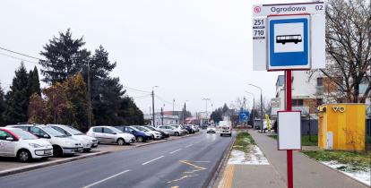 Ulica. Po prawej stronie znajduje się chodnik. Na nim stoi wysoki słup z tabliczką – nazwą przystanku autobusowego i rozkładem jazdy. 