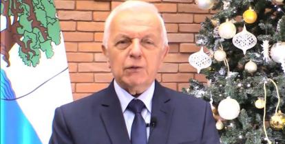 Mężczyzna o siwych włosach w garniturze (burmistrz) na tle ściany z czerwonej cegły, choinki i flagi Konstancina-Jeziorny.