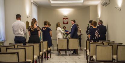 Sala ślubów cywilnych. Na środku naprzeciwko siebie stoi para młoda – trzymają się za ręce, składają przysięgę. Po bokach sali stoją goście – około 10 osób. 