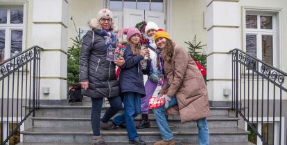 Na schodach przed budynkiem stoją cztery osoby z puszkami WOŚP oraz nalepkami – czerwonymi serduszkami WOŚP.