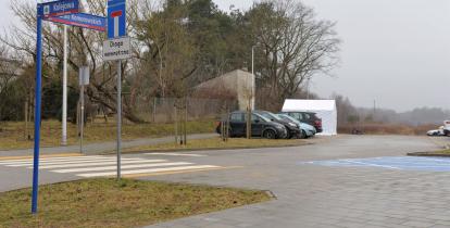 Parking z kostki brukowej. Poi lewej stronie widoczny drogowskaz z nazwami ulic. W tle kilka zaparkowanych samochodów. Na końcu parkingu stoi biały duży namiot. 