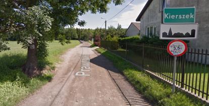 Polna droga, po prawej stronie znak drogowy z nazwą miejscowości Kierszek i zabudowania mieszkalne i gospodarcze, w tle drzewa.