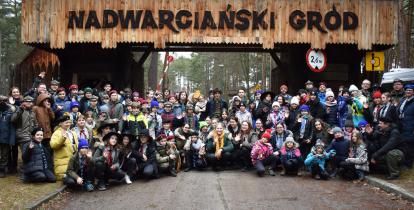 Grupa kilkuset osób w strojach harcerskich na tle drewnianej bramy wjazdowej z napisem: Nadwarciański Gród.