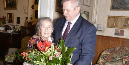 Stojący po prawej stronie mężczyzna w średnim wieku, ubrany w garnitur, wręcza bukiet kwiatów starszej kobiecie stojącej po lewej stronie. Kobieta patrzy na wprost. Za nimi widać mieszkanie, ze ścianami udekorowanymi licznymi obrazami.