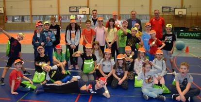 Grupa około 30 dzieci w wieku szkolnym sto w trzech rzędach na sali sportowej. Są ubrani na sportowo, na głowie mają pomarańczowe czapki z daszkiem, a w rękach trzymają zielone odblaskowe sportowe torby. Za nimi stoi 4 dorosłych mężczyzn. 