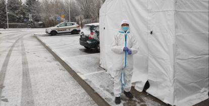 Parking samochodowy z kostki brukowej. Po prawej stronie widoczny biały duży namiot, przed którym stoi osoba w białym kombinezonie i maseczce, z tyłu stoją dwa samochody osobowe.