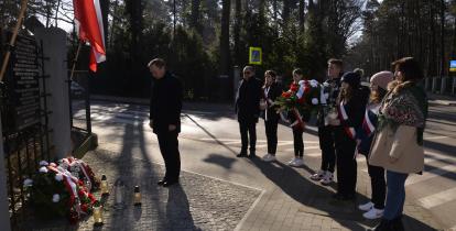 Zdjęcie przedstawia sytuację opisaną w artykule. Na pierwszym planie stoi mężczyzna z głową pochyloną w stronę lewą, gdzie widoczny jest fragment tablicy pamiątkowej, przed tablicą leżą wiązanki kwiatów w kolorach biało-czerwonych oraz znicze, po prawej stronie w rzędzie stoi piątka młodzieży z szarfami biało czerwonymi przepasanymi przez piersi, jeden chłopak trzyma wiązankę kwiatów, po lewej stronie rzędu stoi drugi mężczyzna, po prawej stronie stoi kobieta.